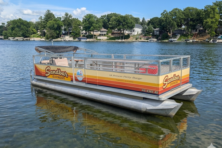 Pontoon boat named 'Sandbar Express' on a lake with trees and houses in the background.