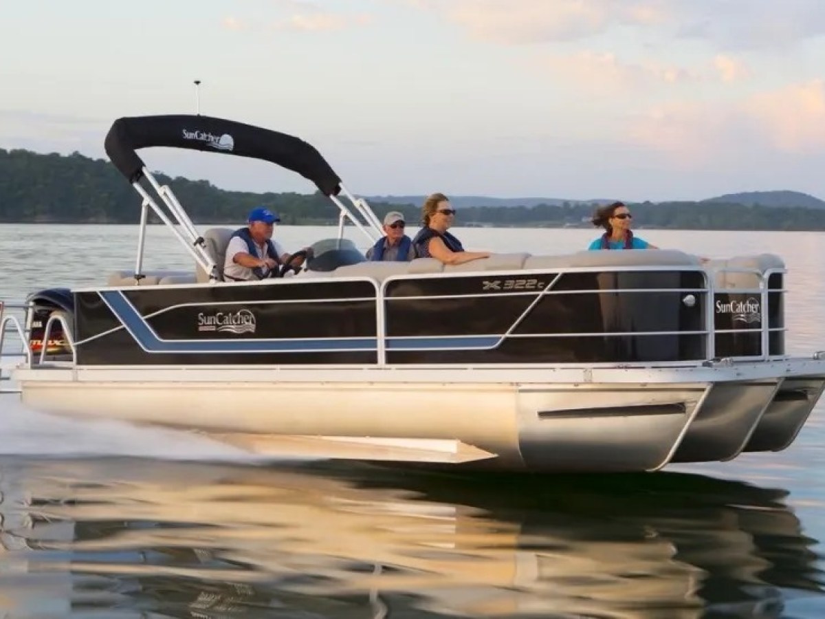 People riding a black pontoon boat on calm water with trees in the background.