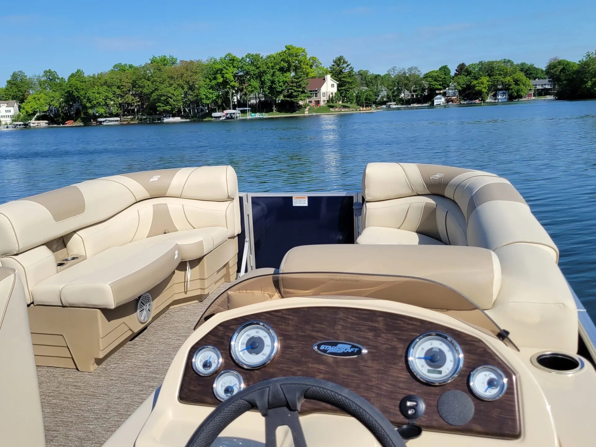 Pontoon boat interior with beige seating on a lake, trees and houses in background.