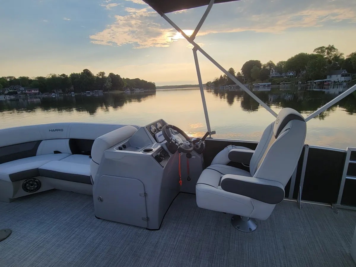 Empty helm of a pontoon boat on a calm lake at sunset with trees and houses in the background.