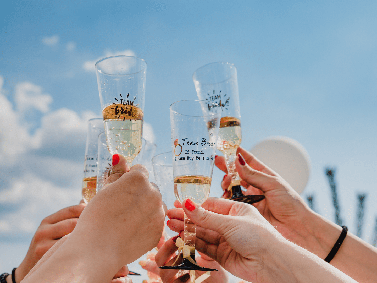 Hands raise champagne glasses with 'Team Bride' under a blue sky.