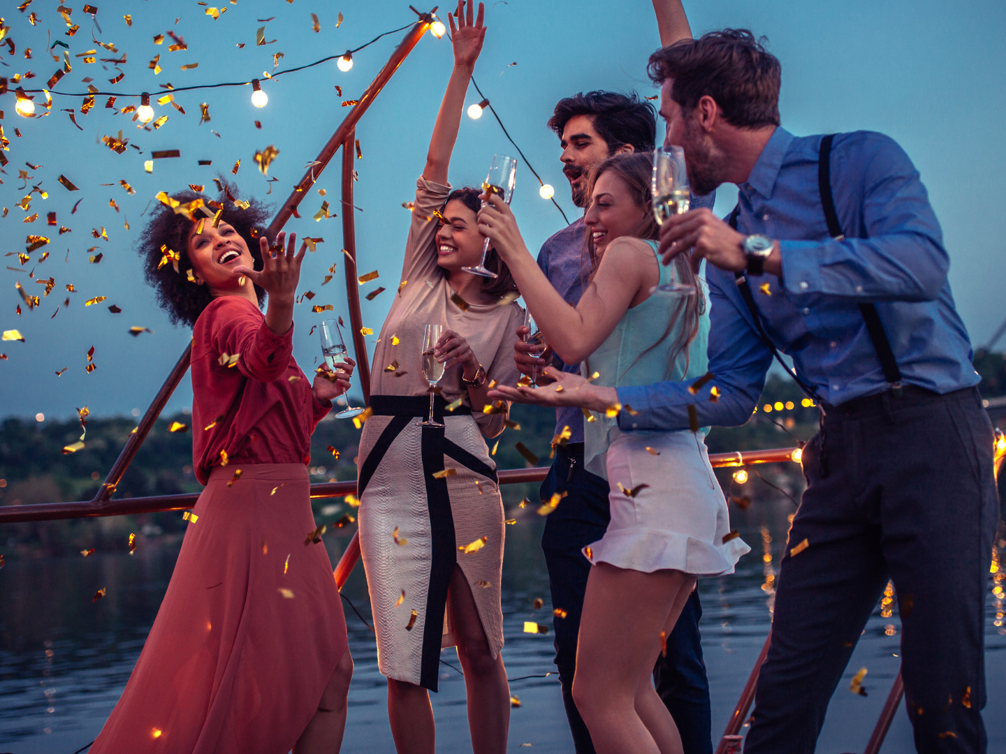 Five people celebrating with confetti and sparklers on a boat with a bridge in the background at dusk.