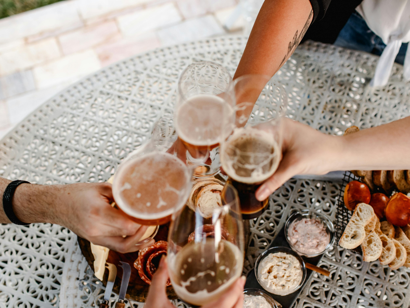 Hands toasting with beers over snacks on a patio table.