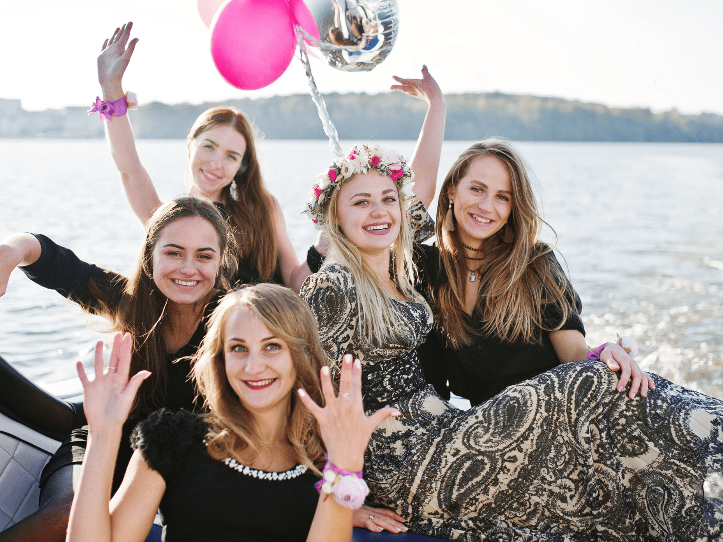 Five women on a boat, smiling and waving, with pink and silver balloons.
