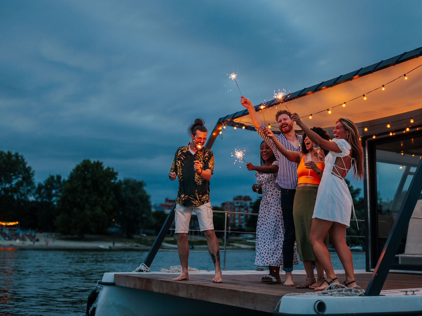 Group of friends with sparklers celebrating on a lit deck at dusk.