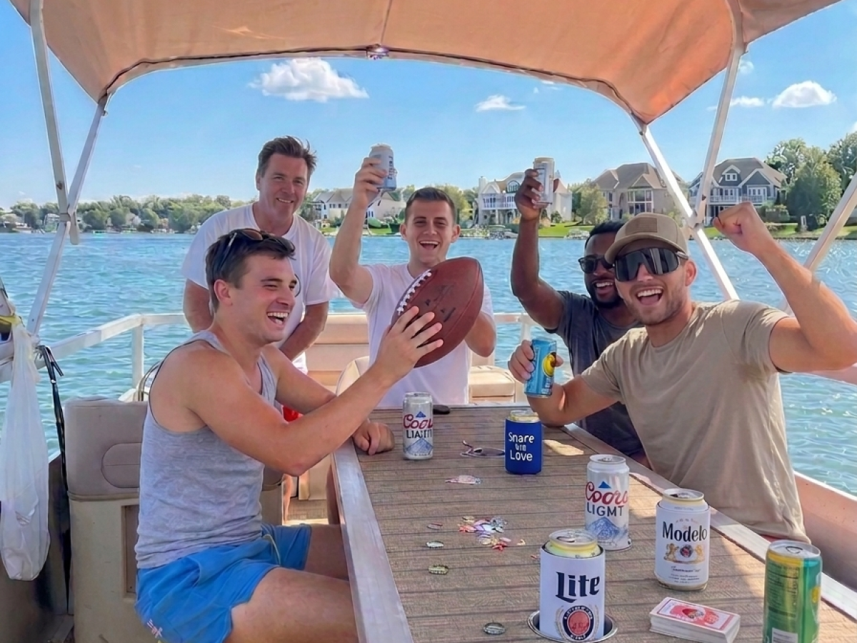 Group of friends on a boat, holding drinks and a football, enjoying a sunny day.